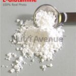 Eine Nahaufnahme zeigt ein weißes, pulverförmiges L-Glutamin-Präparat auf einer grauen Oberfläche. Ein Messlöffel aus Metall entnimmt etwas davon. Der Schriftzug „L-Glutamin“ in Rot und ein Wasserzeichen von „NutriAvenue“ sind erkennbar. 